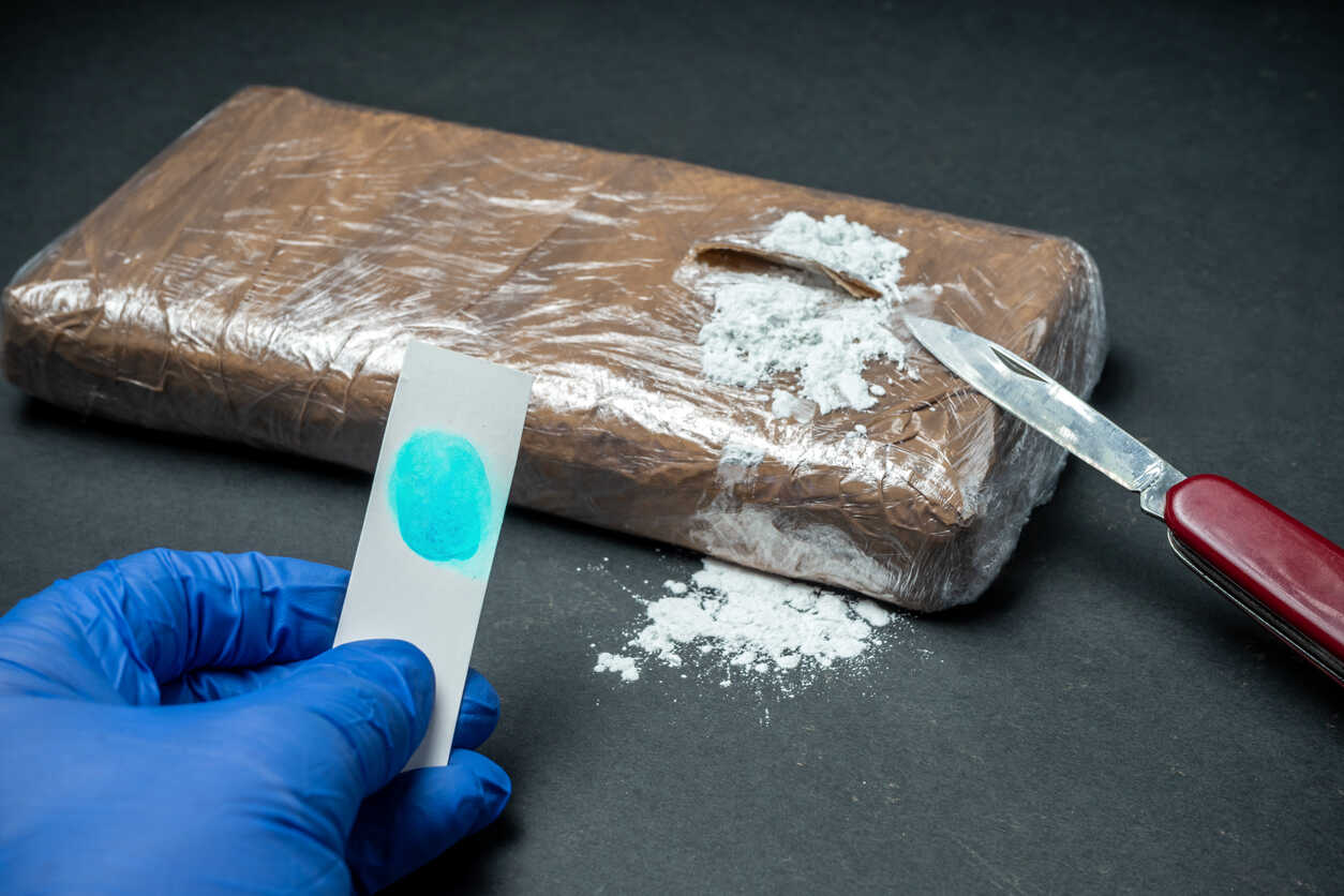 Drug Trafficking A gloved hand holds a test strip next to a large block of a substance and a knife. The image represents a drug trafficking investigation, illustrating the serious nature of the crime and Georgia's laws on the matter.